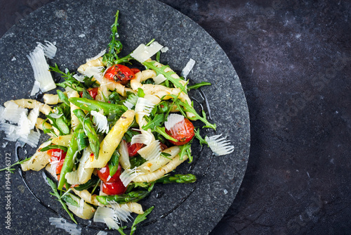 Cold vegetarian pasta noodle salad with green and white Asparagus tips, arugula, fresh tomatos and parmesan served as top view on a black stone plate