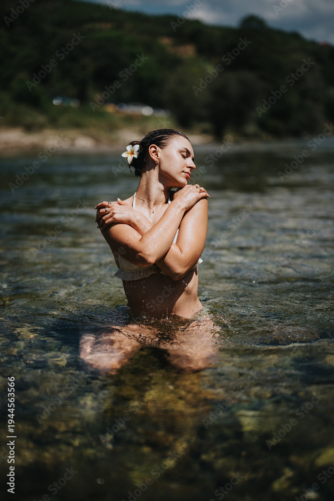 Fototapeta premium A woman stands in clear river water, arms crossed over her chest, eyes closed. The sunlit scene captures a moment of calm and natural beauty by the river.