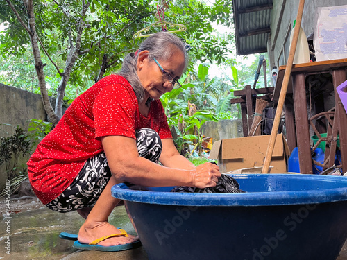 Senior woman hand washing clothes in large blue basin on outdoor patio during rain, elderly lady doing domestic laundry chores at home