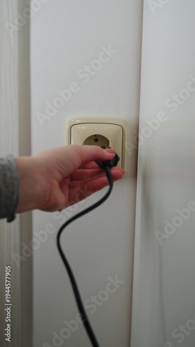Close-up of hand trying to plug electrical cord into wall outlet, but plug doesn’t fit, showing electrical installation and household interior.