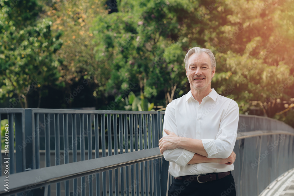 Fototapeta premium Smiling mature man standing with arms crossed near metal railing outdoors with green trees in background and warm sunlight creating peaceful and confident mood