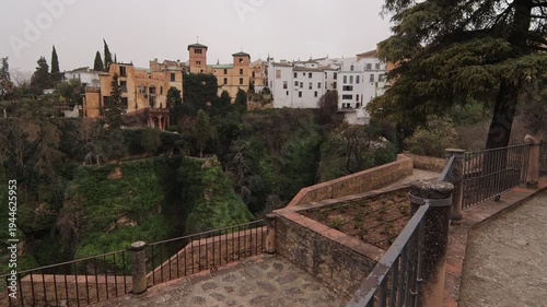 A smooth panning shot showcasing the historic white buildings of Ronda perched on the massive cliffs of El Tajo. Includes the lush vegetation of the gorge and traditional stone pathways.
