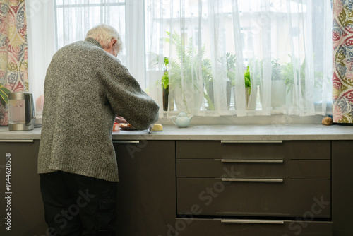 From behind, an elderly man stands at kitchen counter preparing food. The view emphasizes his posture and quiet focus of daily chores. Grandpa engaged in cooking.
