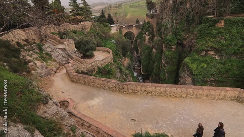 High angle shot of a stone walkway in Ronda, Malaga, featuring two people walking. The scene opens up to the dramatic El Tajo canyon with the historic Puente Viejo bridge in the background.