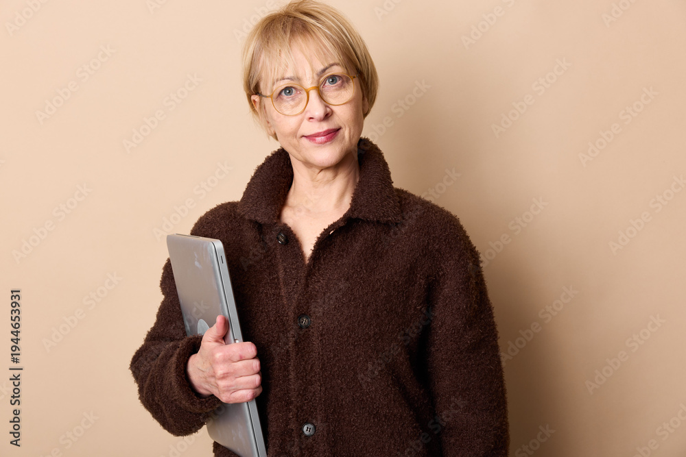 Naklejka premium Mature woman with glasses wearing a brown jacket holds a laptop and looks at the camera. Neutral background with soft lighting creates a calm professional atmosphere.