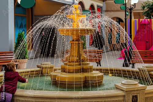 Little girl looking at decorative water fountain in shopping mall. Child fascinated by flowing water stream in public space.