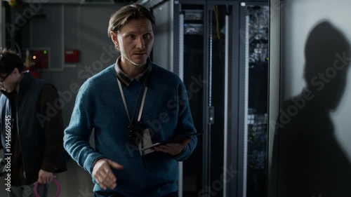 Medium tracking shot of Caucasian male data center technician checking server rack connections while using tablet for system monitoring inside modern server facility
