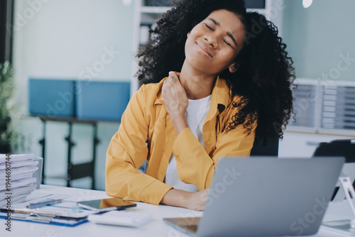 Businesswoman experiencing discomfort and pain in her lower back while sitting at her desk, working on a laptop in a modern office environment, highlighting work-related health issues and stress