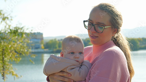 A loving mother in a pink sweater and glasses gently holds her baby boy. They are outdoors by a calm lake, with soft, natural sunlight creating a warm and peaceful mood. Close-up composition