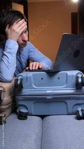 Vertical video stressed male traveler looking at a laptop while sitting with suitcases, experiencing difficulties and problems with flight booking or vacation planning