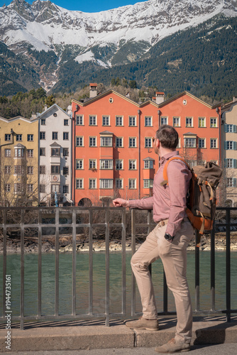 Adventurous man gazes from bridge over Inn river toward vibrant colorful houses of Mariahilfstraße in Innsbruck with alpine peaks, authentic travel vista ideal for lifestyle campaigns.