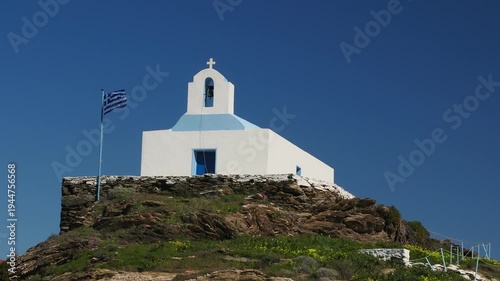 Traditional white Greek island chapel on a rocky hill on Kea Island in the Cyclades, Greece, with the Greek flag fluttering strongly in the wind and natural sea and wind sound.