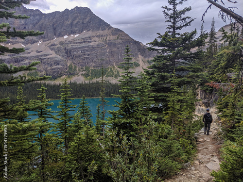 A man hikes on a rocky trail in a forest beside a turquoise lake and a tall mountain in the Canadian Rockies during summer.