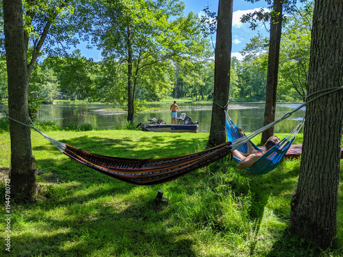 Two colourful hammocks are tied to trees over a grassy shore by a river. One man rests in a hammock while another fishes on his boat near the shoreline.
