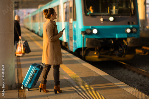 Standing on a sunny railway platform, a woman in a tan coat manages her travel plans on a smartphone, with a blue suitcase and modern train behind her.