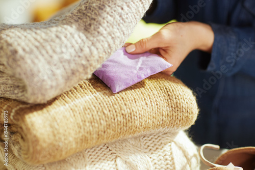 Close-up shot of hands carefully tucking a lavender sachet into a stack of folded knitted sweaters to protect against moths and maintain freshness during storage.