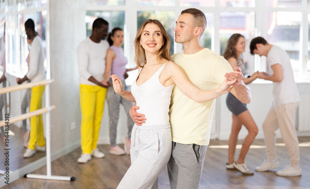 Fototapeta premium Smiling young adult man and woman in casual sportswear enjoying impassioned merengue at Latin dance class in sunlit studio. Social dancing concept..