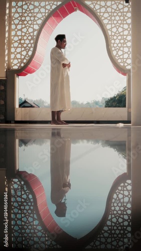 Muslim man performing Islamic prayer inside mosque, peaceful Ramadan worship moment with beautiful arch window and reflection.