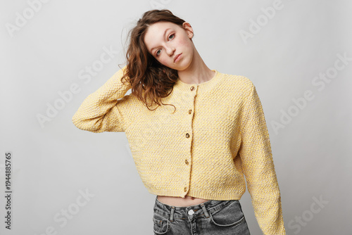 Young woman in yellow knitted cardigan and grey jeans poses with hand in hair against plain light background. Casual fashion, portrait, relaxed mood, natural beauty, lifestyle concept.