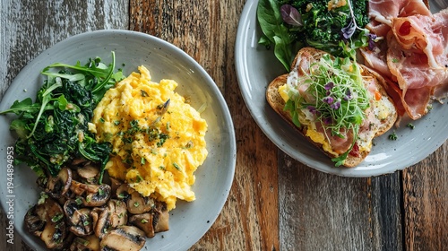 Two people's breakfast plates, viewed from above, feature scrambled eggs, leafy greens, ham, and a cream cheese sandwich with fried mushrooms.