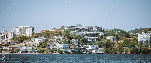 Telephoto panorama of Niteroi waterfront with modern hillside apartments, luxury residences and lush coastal greenery rising above calm bay under clear blue sky, upscale urban shoreline scene