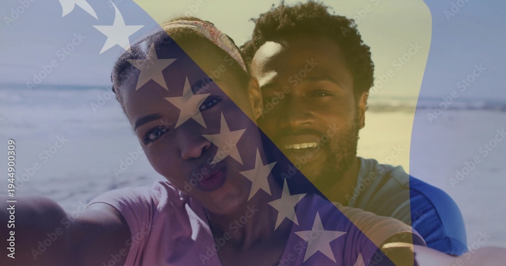 Fototapeta premium Posing couple wearing T-shirts, puckering lips while leaning in for selfie at beach, flag overlay