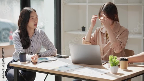 Two young Asian business professionals are sharing a laptop while sitting in a meeting, analyzing financial reports, using a calculator, and discussing data on charts.