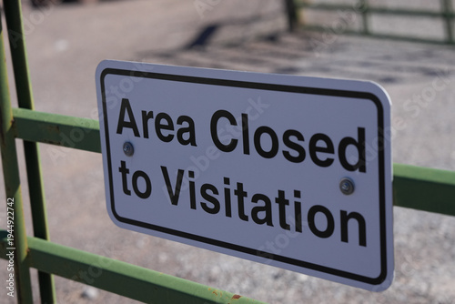 White rectangular “Area Closed to Visitation” sign is bolted to a green metal fence with a dry desert landscape and blue sky softly blurred in the background.