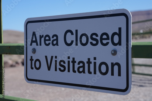 White rectangular “Area Closed to Visitation” sign is bolted to a green metal fence with a dry desert landscape and blue sky softly blurred in the background.