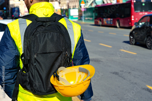 Worker walks on street carrying backpack and hard hat during daytime in a busy city
