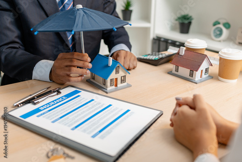 A real estate agent explains home insurance coverage and cost options to a client, holding an umbrella over a house model to symbolize property protection and financial security in real estate.