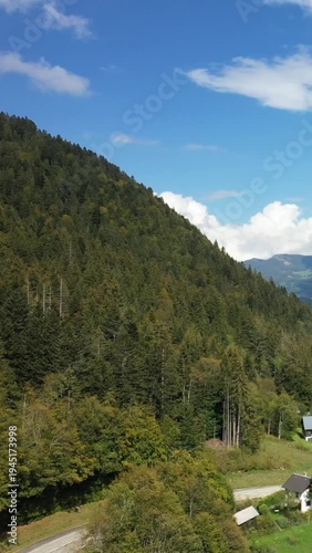 Lush green forest covering a hillside in the Massif de la Chartreuse under a blue sky with scattered clouds. A few rural houses are visible at the forest edge.