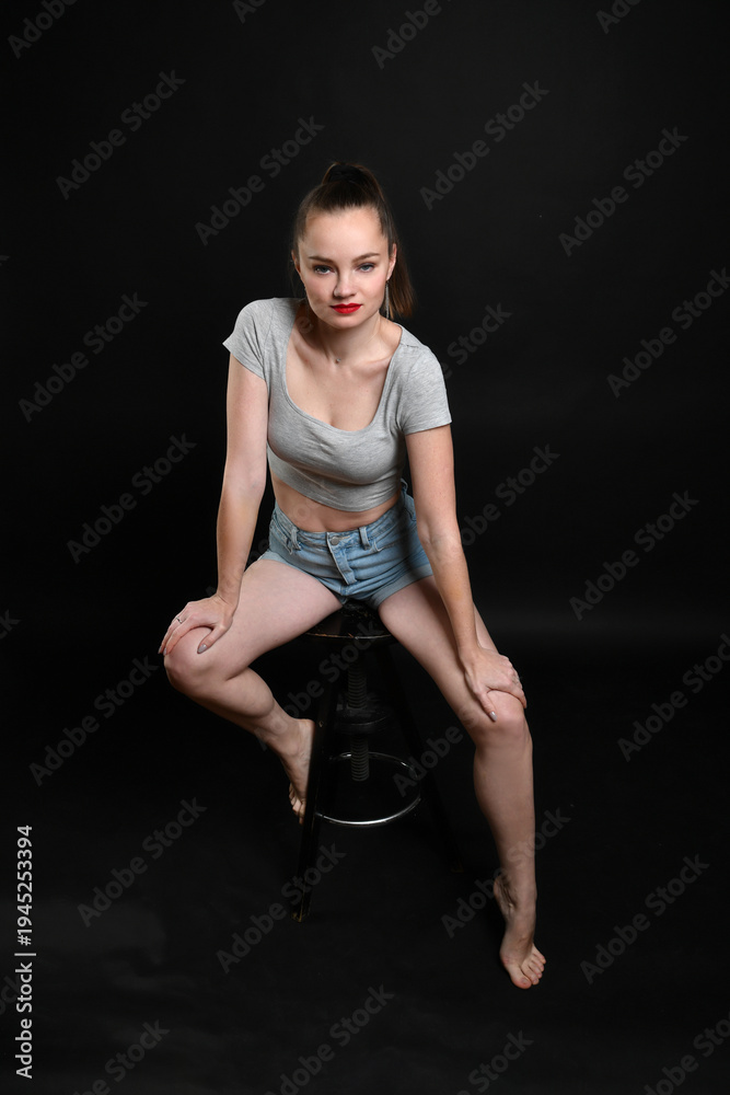 Fototapeta premium Selbstbewusstes Model sitzt auf einem Hocker im Studio und blickt direkt in die Kamera. Modernes Fashion-Porträt mit natürlichem Stil, Konzept für Mode, Lifestyle und Persönlichkeit.