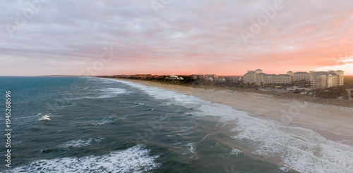 Aerial view of a serene coastal scene where the ocean's foamy waves meet the sandy beach, overlooked by buildings under a pastel-colored sky, Amelia Island, Florida, United States.