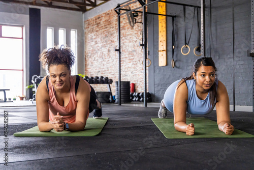 Diverse female workout partners in tanks holding forearm plank on green mats in gym weights visible
