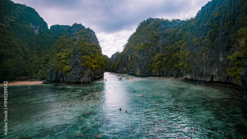 Aerial view of azure waters meet dramatic limestone cliffs, a hidden cove beckons with its sandy shore and scattered boats, El Nido, Mimaropa, Philippines.