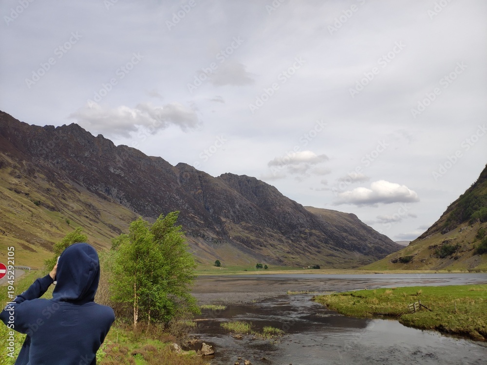 Fototapeta premium Person taking photo of mountain valley landscape with river and cloudy sky in Scotland