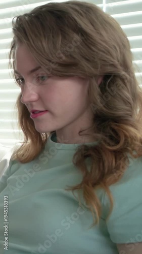 Vertical video: Turning woman in light green tee glancing left as blue arm showing on sofa, blinds
