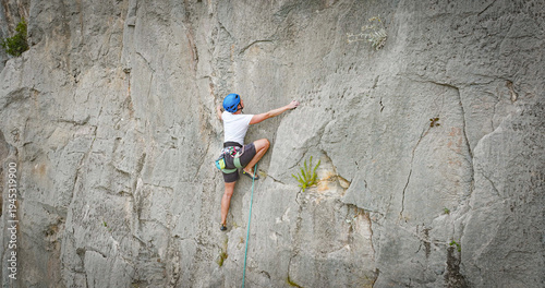 AERIAL, CLOSE UP: Sport climber stretches for next handhold on a steep limestone wall, wearing a helmet and harness with rope running below. Concentration and agile movement in rugged natural setting.