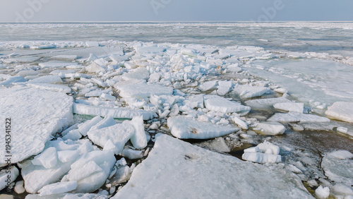 Ice floes and fragmented ice sheets scattered across a frozen lake surface under a clear sky, with distant horizon blending into the icy landscape