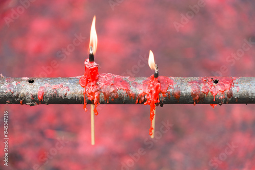 Two vibrant red candles are burning on a weathered metal rod, with melted wax dripping down the sides. The background is a blurred, fiery red, creating a dramatic and intense atmosphere. 