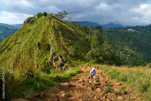 Solo female traveler conquering steep, rugged slope of Little Adam's Peak during golden hour, with majestic Adam's Peak mountain range glowing in warm sunset light, Ella, Sri Lanka.