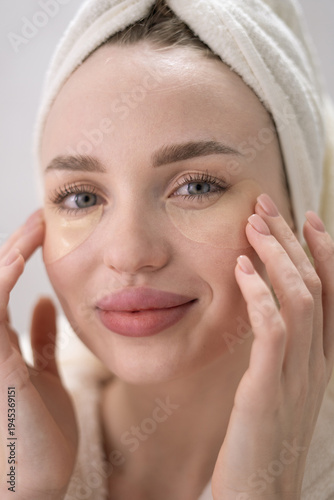 Portrait of Young woman with towel on head after showering applying under eye patches, treatment dark circles at home. Girl using cosmetic products under her eyes.