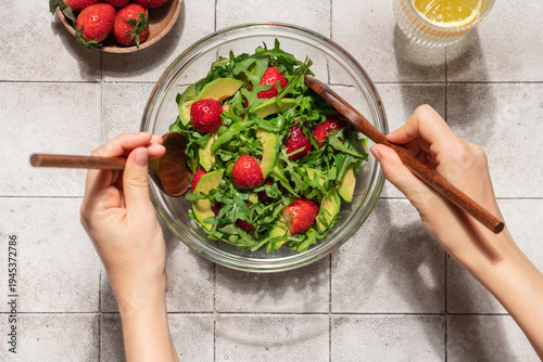 Female hands mixing fresh summer salad with avocado arugula and strawberries in glass bowl on gray tile table, top view healthy homemade meal concept