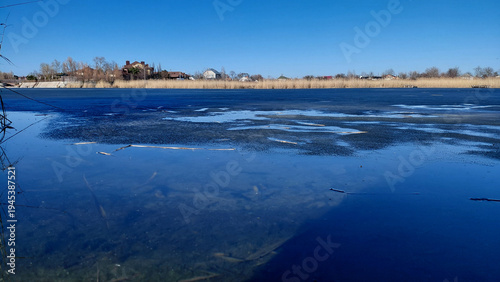 Early spring landscape with a frozen river or lake, clear blue sky reflected in the melt water, and dry reeds on the shore. Ukraine, Dnipro
