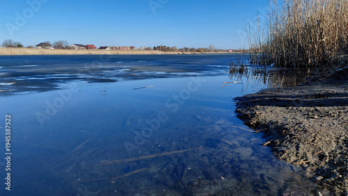 Early spring landscape with a frozen river or lake, clear blue sky reflected in the melt water, and dry reeds on the shore. Ukraine, Dnipro