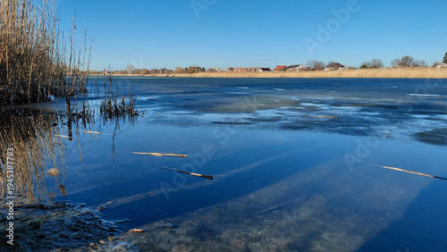 Early spring landscape with a frozen river or lake, clear blue sky reflected in the melt water, and dry reeds on the shore. Ukraine, Dnipro