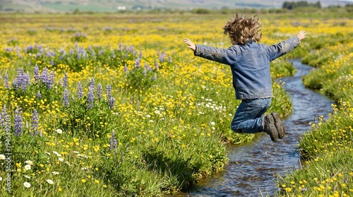 Child jumping over a small stream in a meadow. Boy playing in a field of yellow and purple flowers during spring. Active kid exploring nature, healthy lifestyle and carefree childhood.