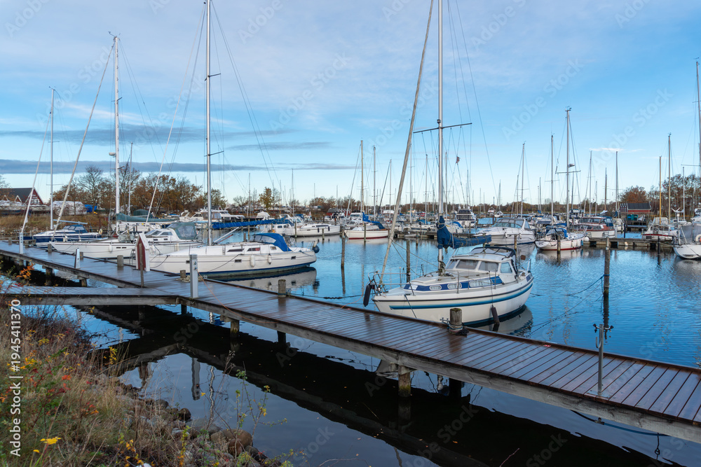 Fototapeta premium Ishoj Marina, Denmark – November 17, 2025: Large marina filled with sailboats and motor yachts moored along floating wooden piers. Tall masts rise above the harbor creating a dense pattern against the