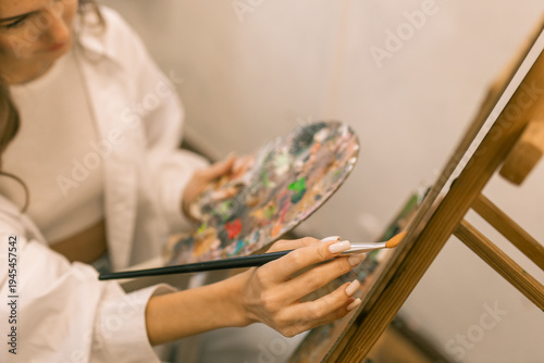 Young woman artist mixing paint on palette while painting on canvas at easel in art studio, creative hobby and painting process
